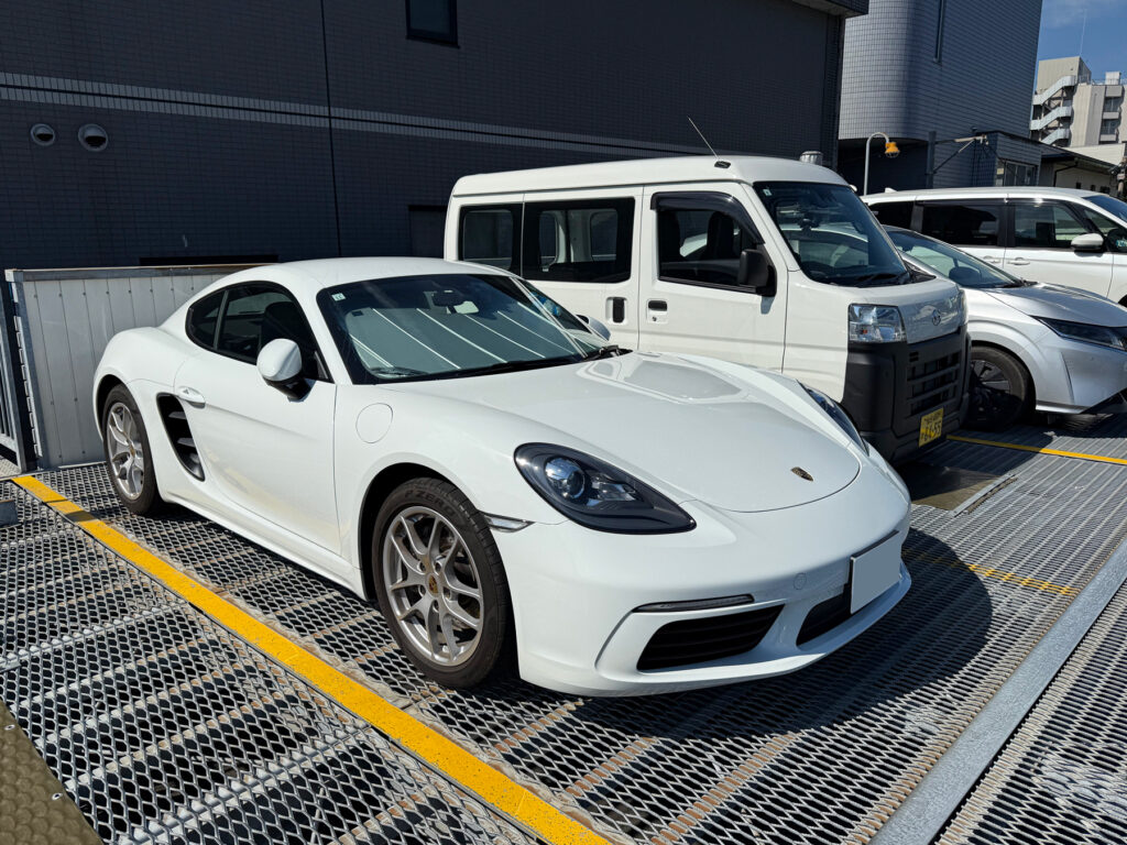 白いポルシェ718ケイマンが駐車場に止まっている様子。周囲には他の車が並び、建物の一部も見える。